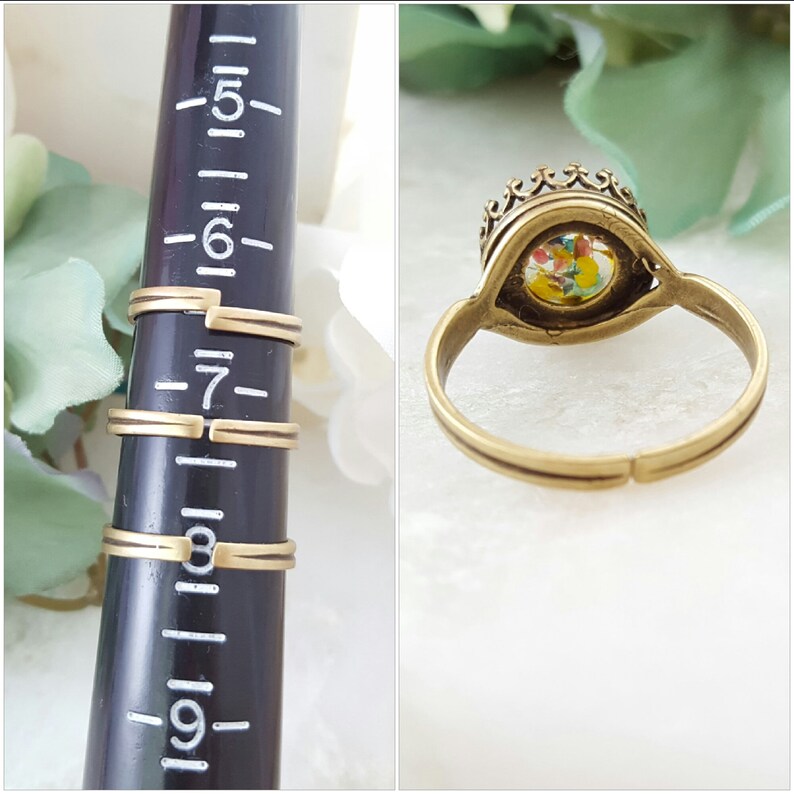 May include: A gold-toned ring with a crown-shaped setting. The setting contains a colorful, speckled stone. The ring is shown on a sizing tool, indicating sizes 5 through 9.