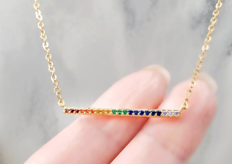 May include: A gold chain necklace with a thin bar pendant set with rainbow colored gemstones.