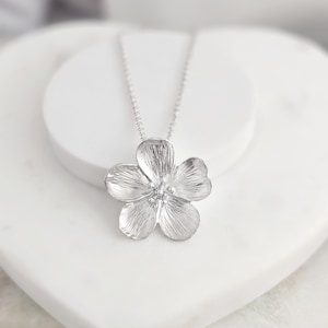May include: A silver flower pendant necklace with a delicate chain. The flower pendant has a textured surface and a small centre stone.