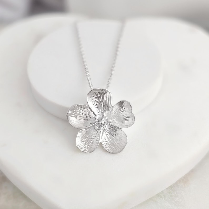 May include: A silver flower pendant necklace with a delicate chain. The flower pendant has a textured surface and a small centre stone.