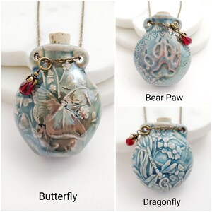 May include: Three ceramic pendant necklaces with different designs: a butterfly, a bear paw, and a dragonfly. Each pendant is a small, blue-green vase shape with a cork stopper, a decorative chain, and a red bead. The pendants hang from a delicate chain.