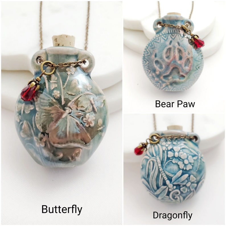 May include: Three ceramic pendant necklaces with different designs: a butterfly, a bear paw, and a dragonfly. Each pendant is a small, blue-green vase shape with a cork stopper, a decorative chain, and a red bead. The pendants hang from a delicate chain.