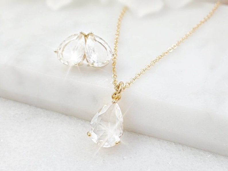 Puede incluir: Un collar de oro con un colgante de cristal en forma de l&aacute;grima y un par de pendientes a juego con l&aacute;grimas de cristal. Las joyas est&aacute;n sobre una superficie blanca.