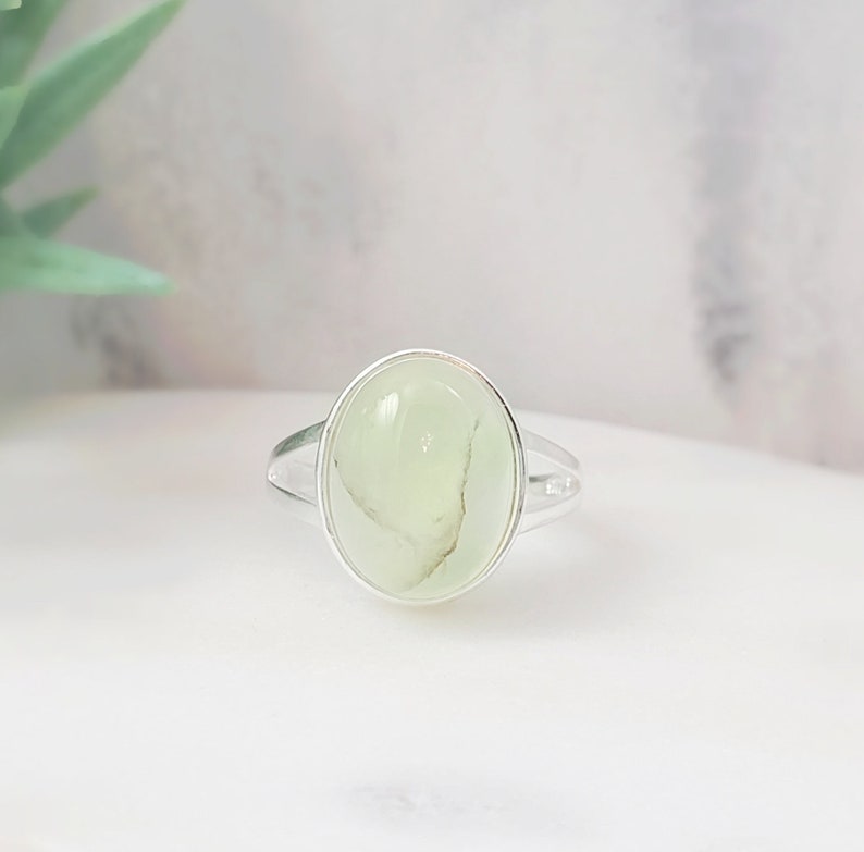 May include: A silver ring with a light green oval-shaped gemstone. The ring is set in a simple, elegant design.