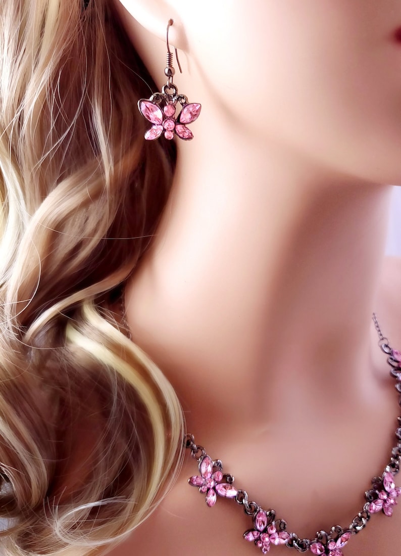 Puede incluir: Un delicado conjunto de collar y pendientes con un dise&ntilde;o de mariposa con detalles de cristal rosa. El collar tiene una cadena con m&uacute;ltiples dijes de mariposa, mientras que los pendientes son de estilo colgante con un solo dije de mariposa.