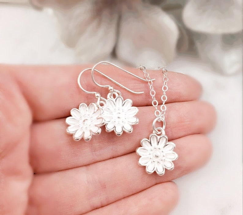 May include: A silver necklace and earrings set featuring a delicate flower design. The necklace has a dainty chain and a small flower pendant. The earrings are also small and feature a flower design.