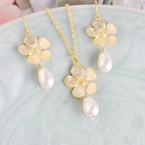 May include: A gold necklace and earrings set featuring delicate flower charms with pearl drops. The flowers have a textured, almost hammered, finish.