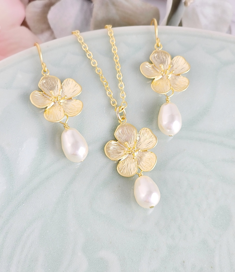 May include: A gold necklace and earrings set featuring delicate flower charms with pearl drops. The flowers have a textured, almost hammered, finish.