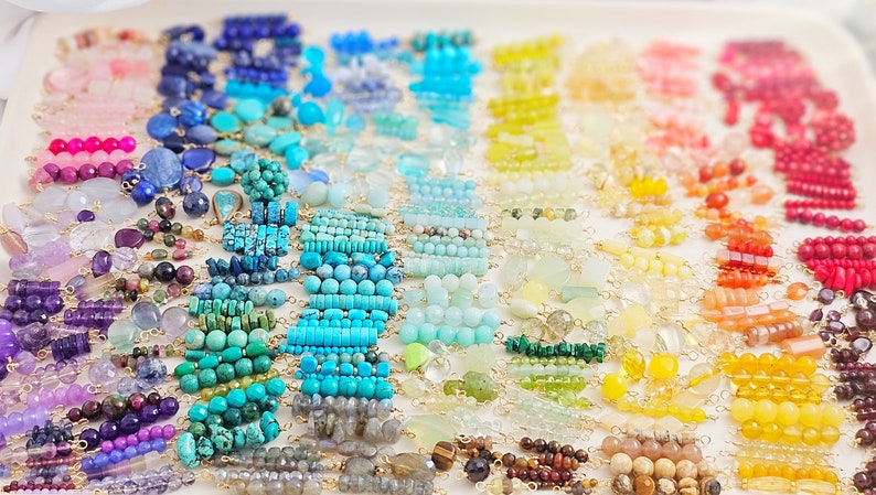 Puede incluir: Un surtido arco&iacute;ris de cuentas de piedras preciosas dispuestas en filas por color. Las cuentas est&aacute;n enhebradas en alambre dorado y est&aacute;n listas para ser utilizadas en la elaboraci&oacute;n de joyas.