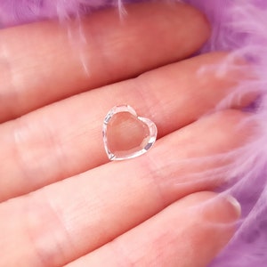 Puede incluir: Un amuleto de cristal transparente en forma de coraz&oacute;n, sostenido en una mano sobre un fondo morado suave.