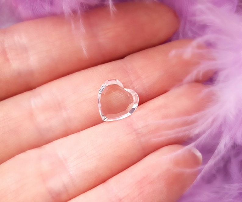 Puede incluir: Un amuleto de cristal transparente en forma de coraz&oacute;n, sostenido en una mano sobre un fondo morado suave.