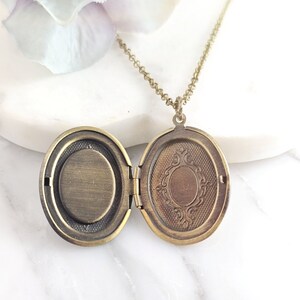 Puede incluir: Un collar con un medall&oacute;n ovalado en una cadena delicada. El medall&oacute;n est&aacute; abierto, revelando un interior liso de color lat&oacute;n. El medall&oacute;n tiene un dise&ntilde;o ornamentado en la parte delantera.