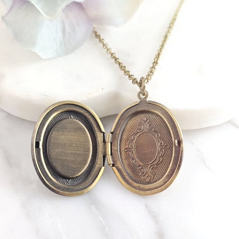 Puede incluir: Un collar con un medall&oacute;n ovalado en una cadena delicada. El medall&oacute;n est&aacute; abierto, revelando un interior liso de color lat&oacute;n. El medall&oacute;n tiene un dise&ntilde;o ornamentado en la parte delantera.