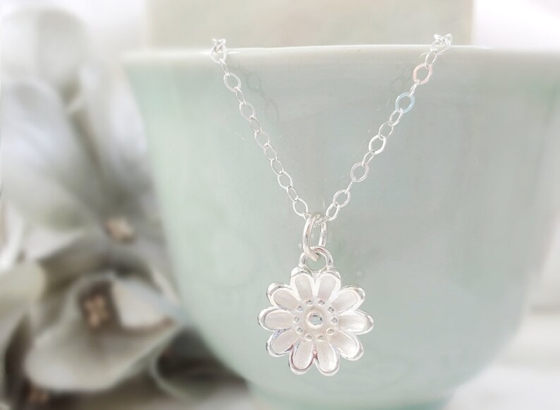 May include: A silver chain necklace with a delicate silver flower pendant. The flower has a textured surface and a small center.