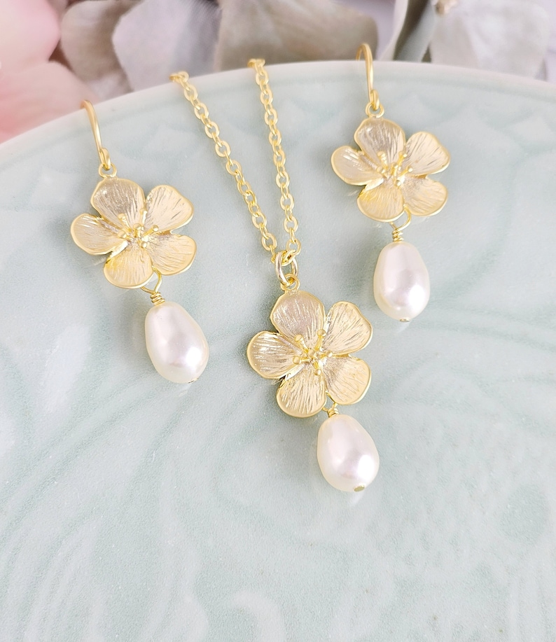 May include: A gold necklace and earrings set featuring delicate flower charms with pearl drops. The flowers have a textured, almost hammered, finish.