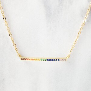 May include: A gold chain necklace with a thin bar pendant encrusted with colorful gemstones. The gemstones are arranged in a rainbow pattern.
