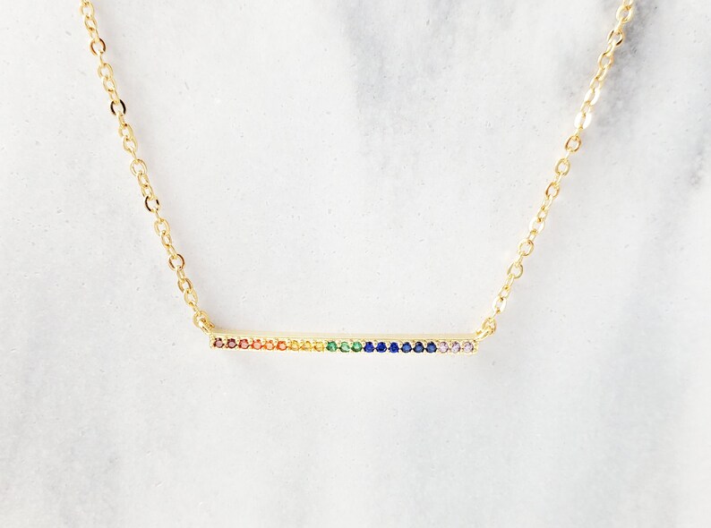 May include: A gold chain necklace with a thin bar pendant encrusted with colorful gemstones. The gemstones are arranged in a rainbow pattern.