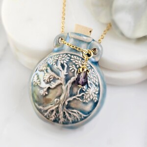 May include: A blue ceramic pendant necklace with a gold chain. The pendant is a bottle shape with a tree design and a cork stopper. A small purple crystal hangs from the chain. The necklace is displayed on a white surface.