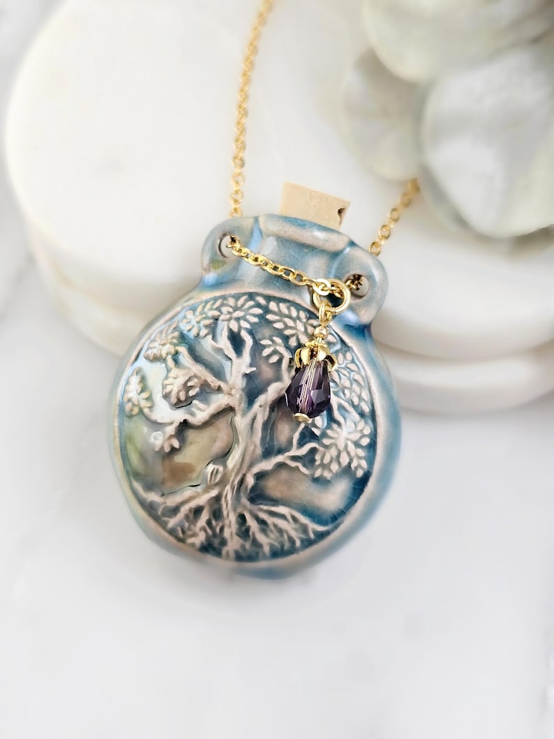 May include: A blue ceramic pendant necklace with a gold chain. The pendant is a bottle shape with a tree design and a cork stopper. A small purple crystal hangs from the chain. The necklace is displayed on a white surface.