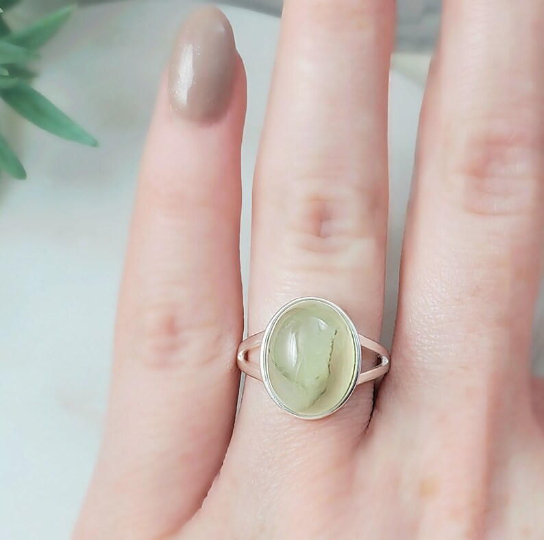 May include: A silver ring with a light green oval gemstone. The ring is on a finger.