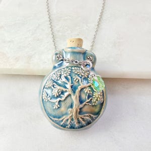 May include: A blue ceramic pendant necklace with a tree of life design. The pendant has a cork stopper and a silver chain. A green crystal charm hangs from the pendant. The necklace is displayed on a white surface.
