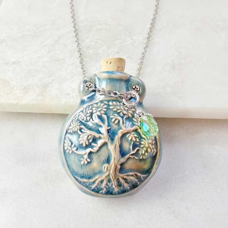 May include: A blue ceramic pendant necklace with a tree of life design. The pendant has a cork stopper and a silver chain. A green crystal charm hangs from the pendant. The necklace is displayed on a white surface.