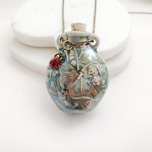 May include: A ceramic pendant necklace featuring a small, blue-green bottle with a cork stopper. The bottle is adorned with a floral and butterfly design, and a red crystal charm hangs from the chain. The necklace is suspended from a bronze-colored chain.