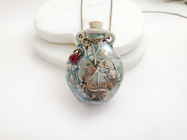 May include: A ceramic pendant necklace featuring a small, blue-green bottle with a cork stopper. The bottle is adorned with a floral and butterfly design, and a red crystal charm hangs from the chain. The necklace is suspended from a bronze-colored chain.