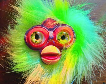 Könnte beinhalten: Ein farbenfrohes, skurriles, Furby-ähnliches Wesen mit großen, ausdrucksstarken Augen und einem lebendigen, federartigen Gefieder. Die Augen sind eine auffällige Kombination aus Rot, Orange und Grün, während die Federn von Gelb zu Grün übergehen. Der Mund ist hellpfirsichfarben.