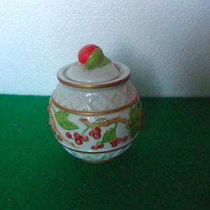 May include: White ceramic jar with a lid decorated with red berries and green leaves. The jar has a gold trim around the top and bottom.