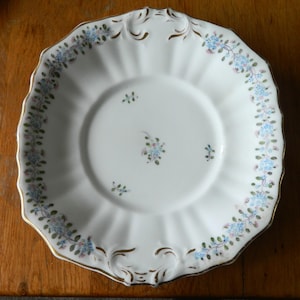 May include: A white porcelain saucer with a scalloped edge and gold trim. The rim features a delicate floral garland of small blue flowers and green leaves. The center of the saucer is decorated with three small, scattered floral sprigs.
