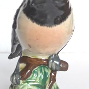 May include: A ceramic bird figurine with a black head, gray wings, and a peach-colored chest. The bird is perched on a green and brown base, holding a small tool. The figurine is a decorative item.
