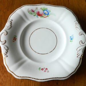 May include: A square white porcelain saucer with rounded corners and scalloped edges. The center features a thin brown circular ring. The rim is decorated with gold trim and small floral motifs including a rose, blue flowers, and pink blossoms.