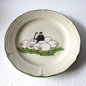 May include: A round ceramic plate with a scalloped edge and a dark green rim. The centre features a colourful illustration of a shepherd in a dark coat holding a wooden staff, surrounded by a small flock of white sheep standing on green grass.