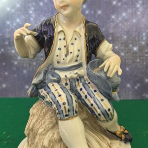 Puede incluir: Figura de porcelana de un niño sentado con un atuendo azul y blanco con detalles dorados. El niño sostiene un sombrero y tiene una expresión serena. La figura está sobre una base decorativa con ribete dorado.