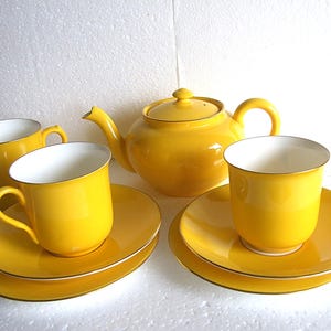 May include: A yellow tea set comprising a teapot, a milk jug, and two cups with saucers. The set has a gold trim. The teapot has a lid and a curved handle. The milk jug has a spout and a handle. The cups and saucers are round.