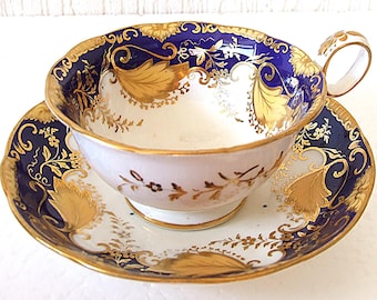 May include: A vintage teacup and saucer set. The cup and saucer are white with a blue border and gold leaf and floral designs. The cup has a handle and the saucer is round.