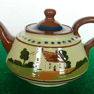 Może przedstawiać: Ceramiczny imbryk z brązowym uchwytem, ​​dzióbkiem i pokrywką. Korpus jest kremowy z namalowaną sceną krajobrazową przedstawiającą dom, drzewa i zieloną trawę. Niebieskie kropki otaczają górną część imbryka.