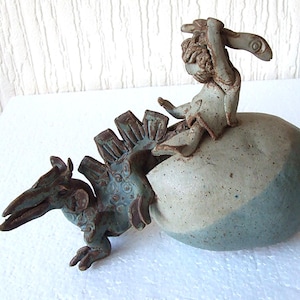 May include: A ceramic sculpture depicting a creature emerging from an egg. The sculpture features a figure on top of the egg, with a detailed, textured design and a color palette of muted greens and browns.
