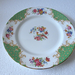 May include: A white porcelain plate with a green and gold patterned border. The plate features a central design of a colourful bird surrounded by floral arrangements in red, pink, blue, and yellow. The plate has a gold rim.