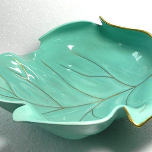 May include: A leaf-shaped ceramic bowl in a bright turquoise colour with a glossy finish. The bowl features delicate gold-coloured trim along the edges and gold-coloured detailing that highlights the veins of the leaf design, including a gold stem.