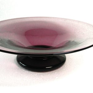 May include: A wide, shallow bowl made of translucent purple glass with a smooth, curved rim. The bowl is supported by a solid, opaque black glass pedestal base that is circular in shape, providing a stable foundation for the vessel.