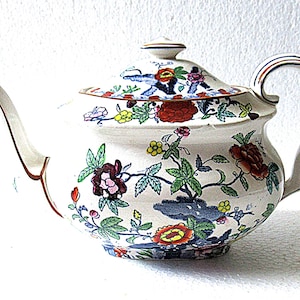 May include: A white ceramic teapot featuring a colourful floral pattern with red, yellow, and blue blossoms, green leaves, and dark blue rock formations. The teapot includes a matching lid with a small knob, a curved handle, and a tapered spout.