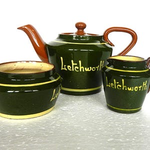 Puede incluir: Un juego de té vintage con un esmalte verde oscuro, que incluye una tetera, una jarra de crema y un azucarero. El juego tiene asas y detalles marrones, con ribetes amarillos y la palabra "Letchworth".