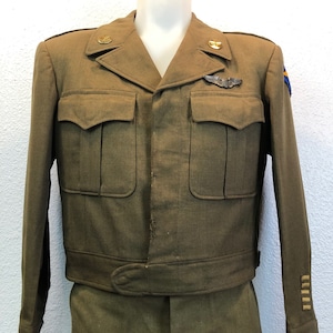 May include: A green military uniform jacket and matching pants. The jacket has a button-up front, two flap pockets, and a belt. The jacket has a silver pilot wing pin and two gold buttons on the collar. The pants have a straight leg and a button fly.