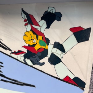 May include: A red, white, and black robot with a yellow head and green accents is in a dynamic pose, appearing to be in mid-air. The robot has a sharp, angular design and is set against a white background.