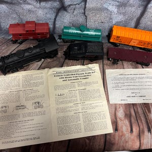 Könnte beinhalten: Ein elektrisches Vintage-Zugset mit einer schwarzen Lokomotive, mehreren bunten Waggons und Bedienungsanleitungen. Die Waggons sind rot, grün, orange, schwarz und weinrot. Die Bedienungsanleitung lautet "Remote Controlled Electric Train Set with Steam Type Locomotive and Automatic Coupler."