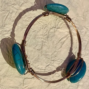 May include: A gold wire bangle bracelet with three turquoise stones. The stones are oval shaped and have a marbled pattern.