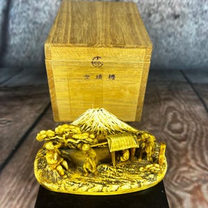 May include: A detailed, golden-colored miniature sculpture depicting a mountain scene with figures, a hut, and trees. The sculpture sits on a black base, with a wooden box in the background. The box has Japanese text and a symbol.