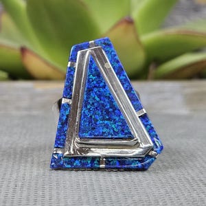 May include: A ring featuring a large, asymmetrical trapezoidal face inlaid with vibrant blue crushed stone. The design incorporates polished silver-toned metal accents that frame the blue inlay, creating a layered, geometric appearance on the surface.